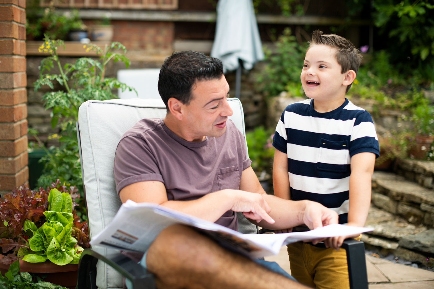 Bild eines Vaters, der Zeitung liest, während sein Sohn mit Down-Syndrom neben ihm lächelt