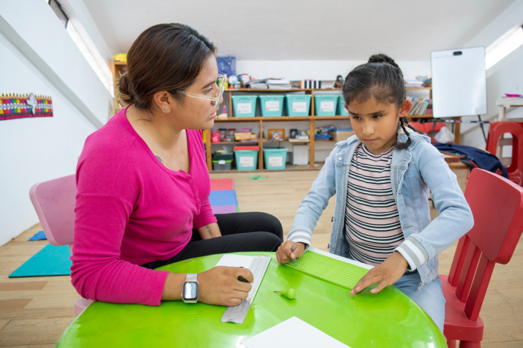 A woman sitting at a desk in a school classroom helping a student that can not see.
