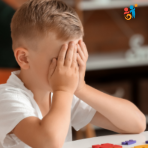 Young boy with blond hair with his hands over his eyes sitting at a desk