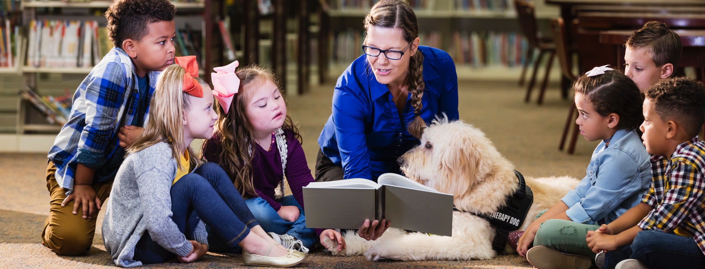 imagen de varios niños sentados en el suelo con el perro de servicio mientras el maestro lee un libro