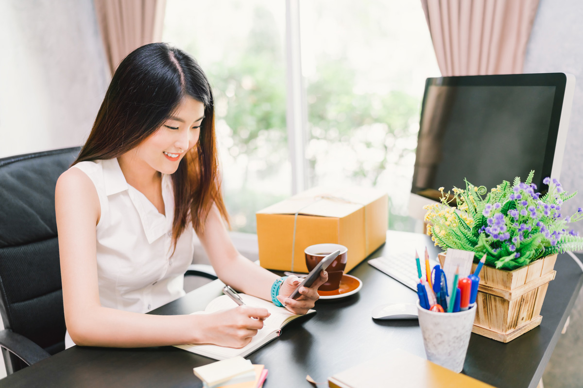 image of female in office chair smiling while making notes and looking at her phone