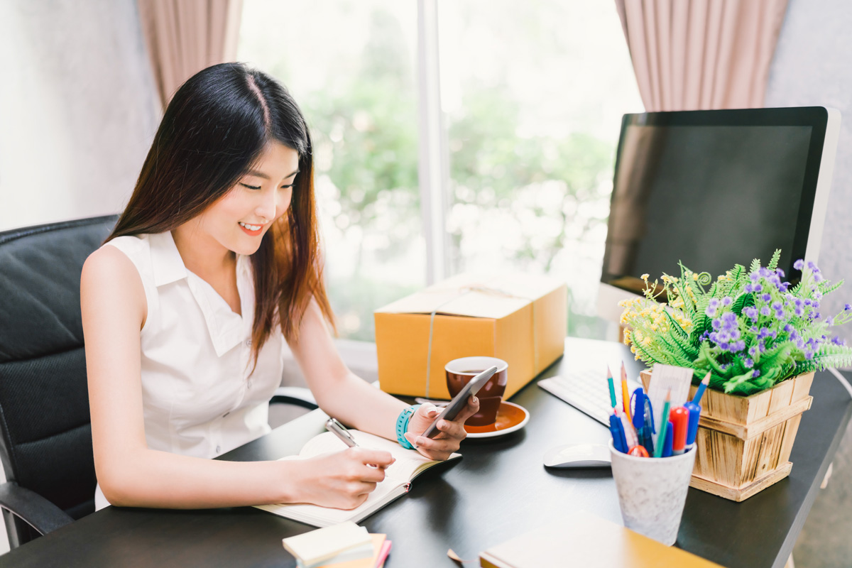 image of female in office chair smiling while making notes and looking at her phone