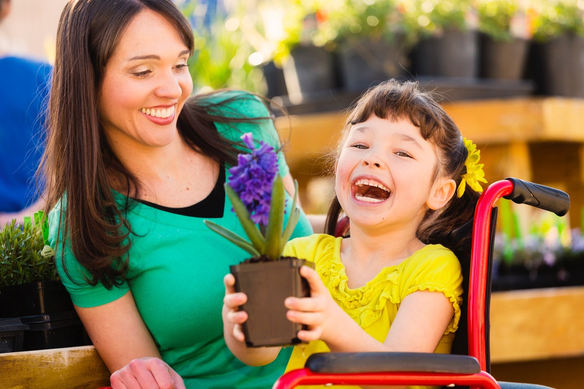 image of young girl in wheelchair holding a plant smiling next to mom