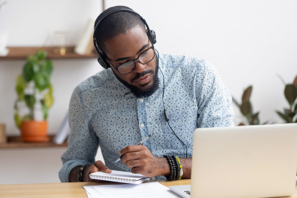 Image of young black male with headphones on taking notes while looking at computer