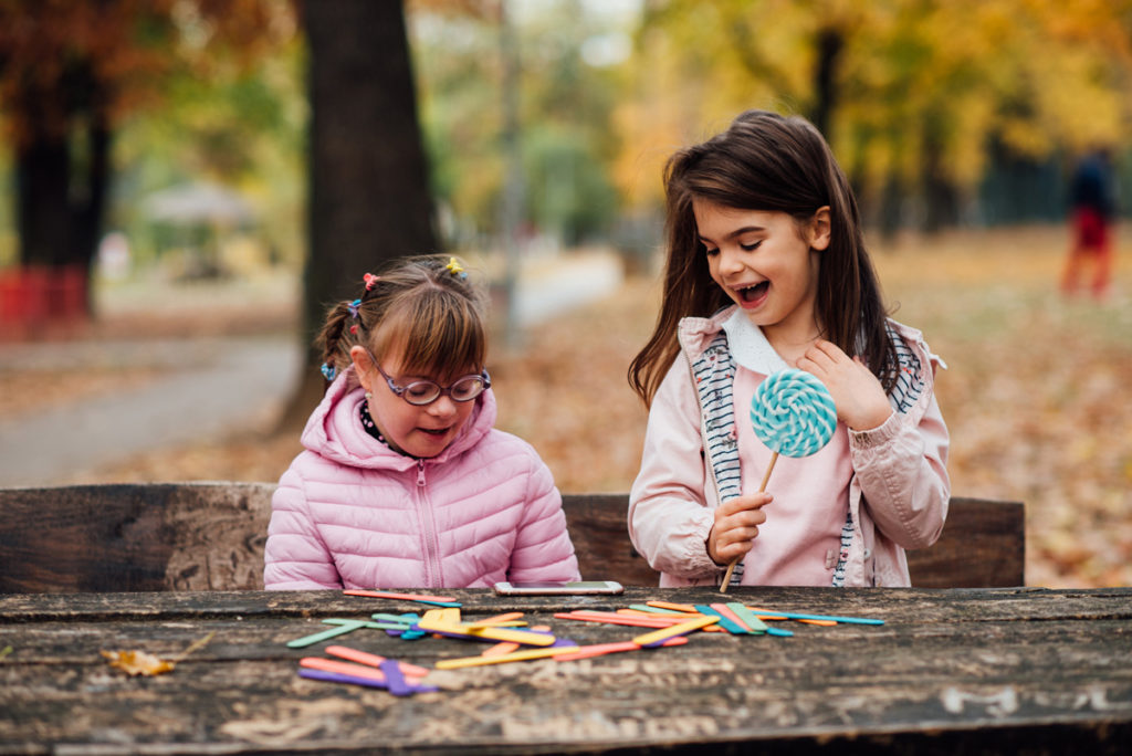 Bild von jungen Mädchen mit Brille draußen spielen mit einem anderen jungen Mädchen hält einen Lolly Pop