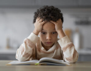 Stressed Little Black Boy Reading Book At Home, Overwhelmed Preteen African American Male Kid Sitting At Desk And Holding Head In Hands, Doing Homework, Suffering Learning Difficulties, Closeup